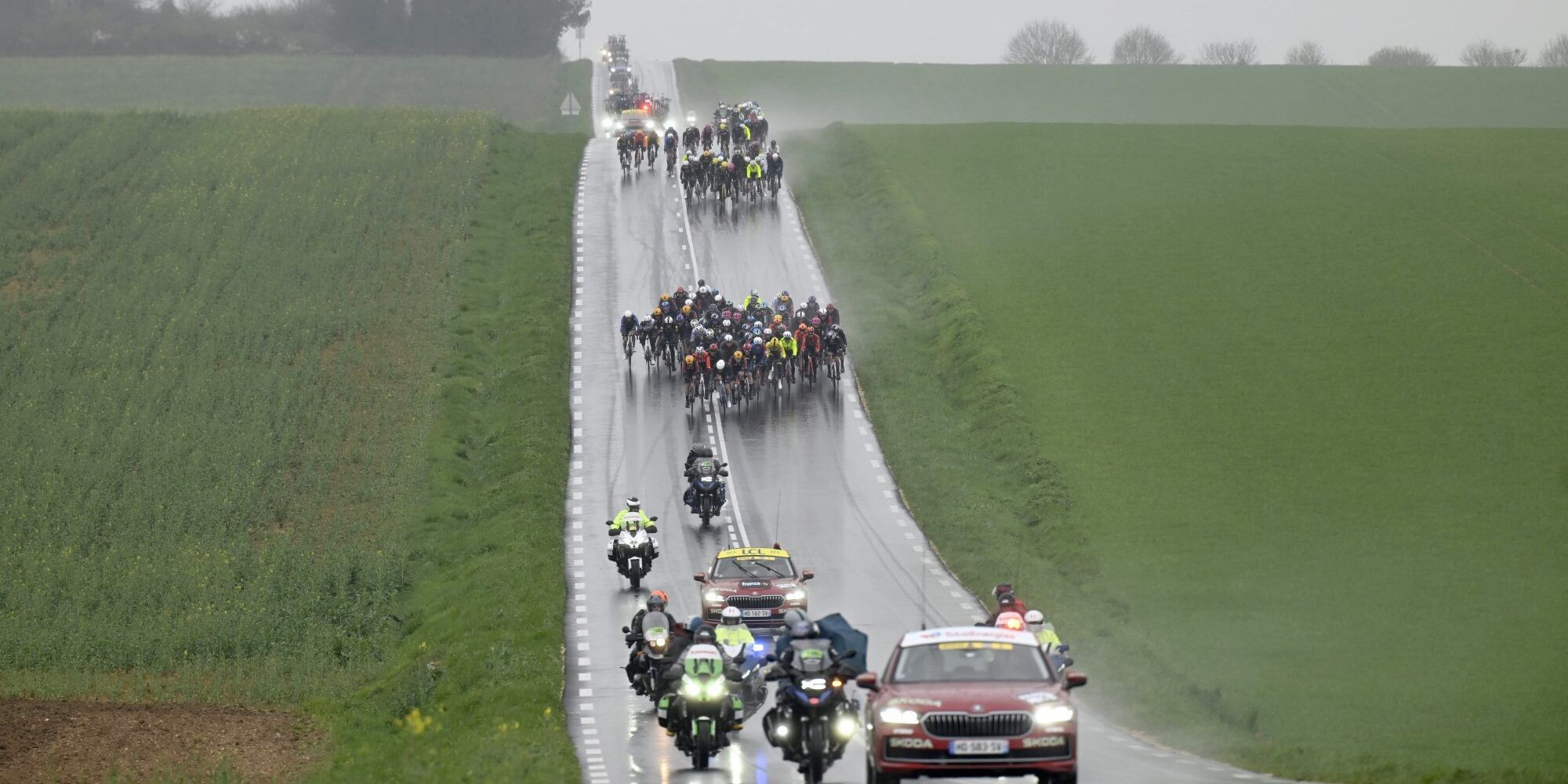 Cycling peloton racing through rain on wet rural road during Paris-Nice stage 4