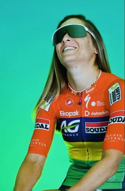 Cyclist in orange AG Insurance-Soudal jersey smiling while wearing sunglasses on turquoise background