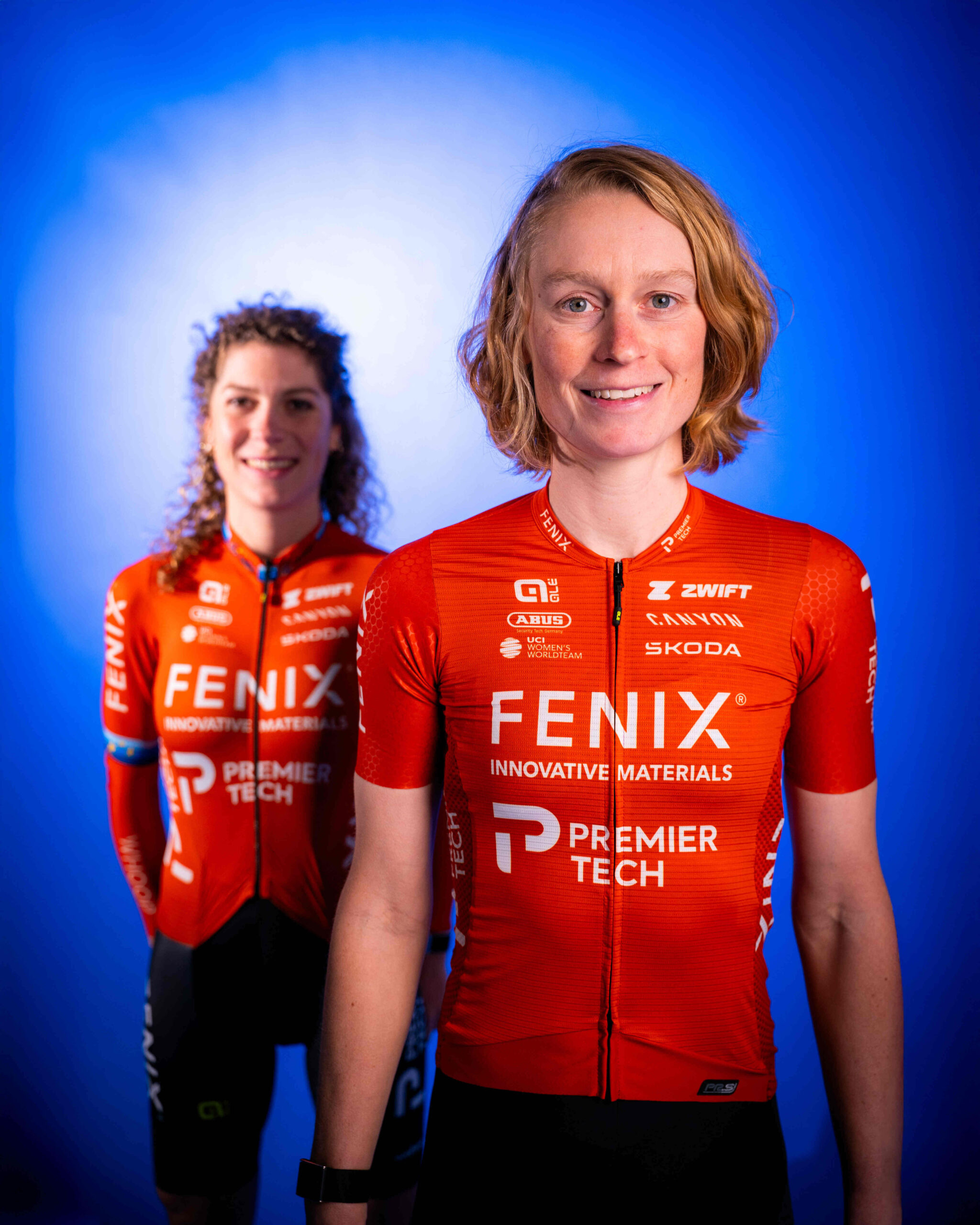 Two female cyclists in red Fenix-Deceuninck team jerseys posing for team portrait against blue background