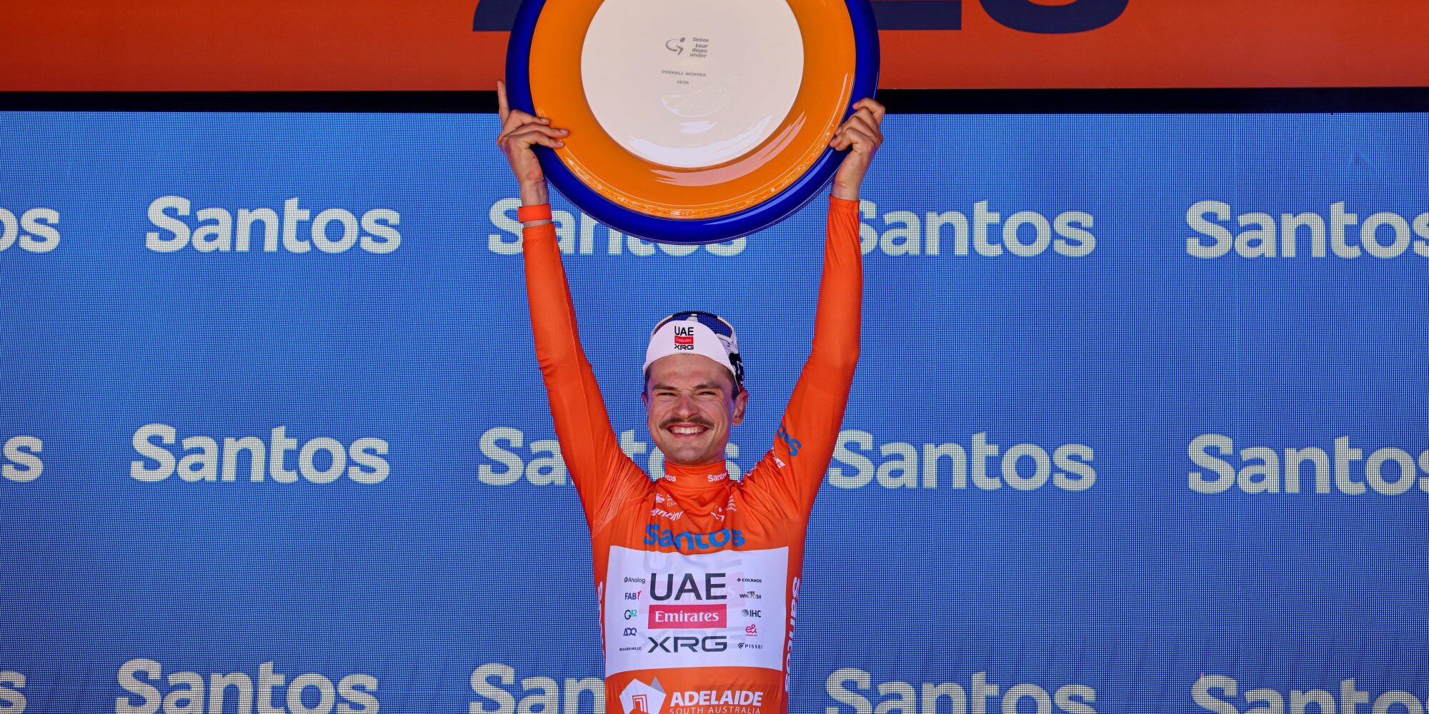 Jay Vine holding orange trophy overhead on Santos Tour Down Under podium in orange leader's jersey